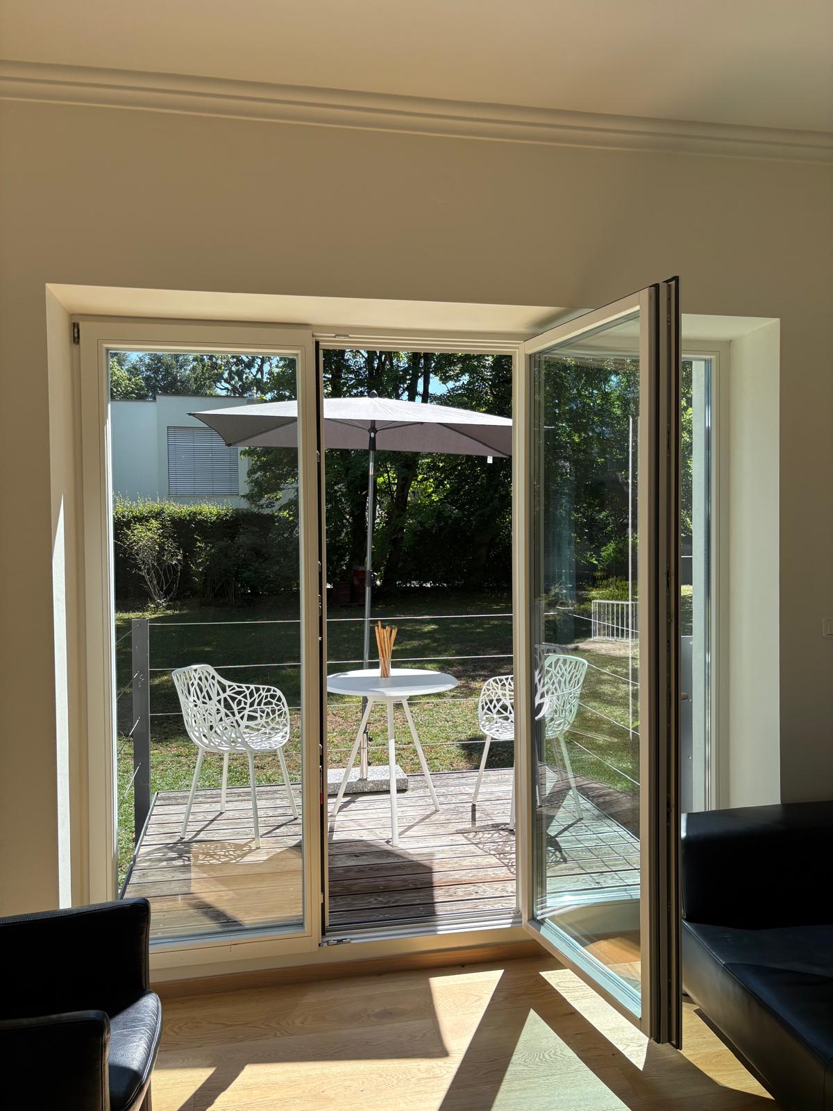Blick durch die geöffnete Fenstertür auf die Terrasse mit Sonnenschirm und Sitzplatz im Grünen
